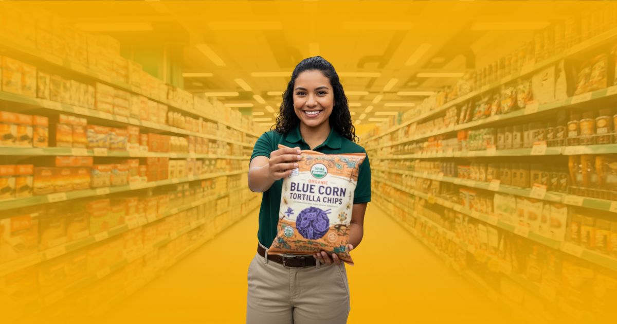 Promotor de ventas sonriente en un stand de degustación de botanas, ofreciendo el producto a una pareja de clientes en un supermercado.